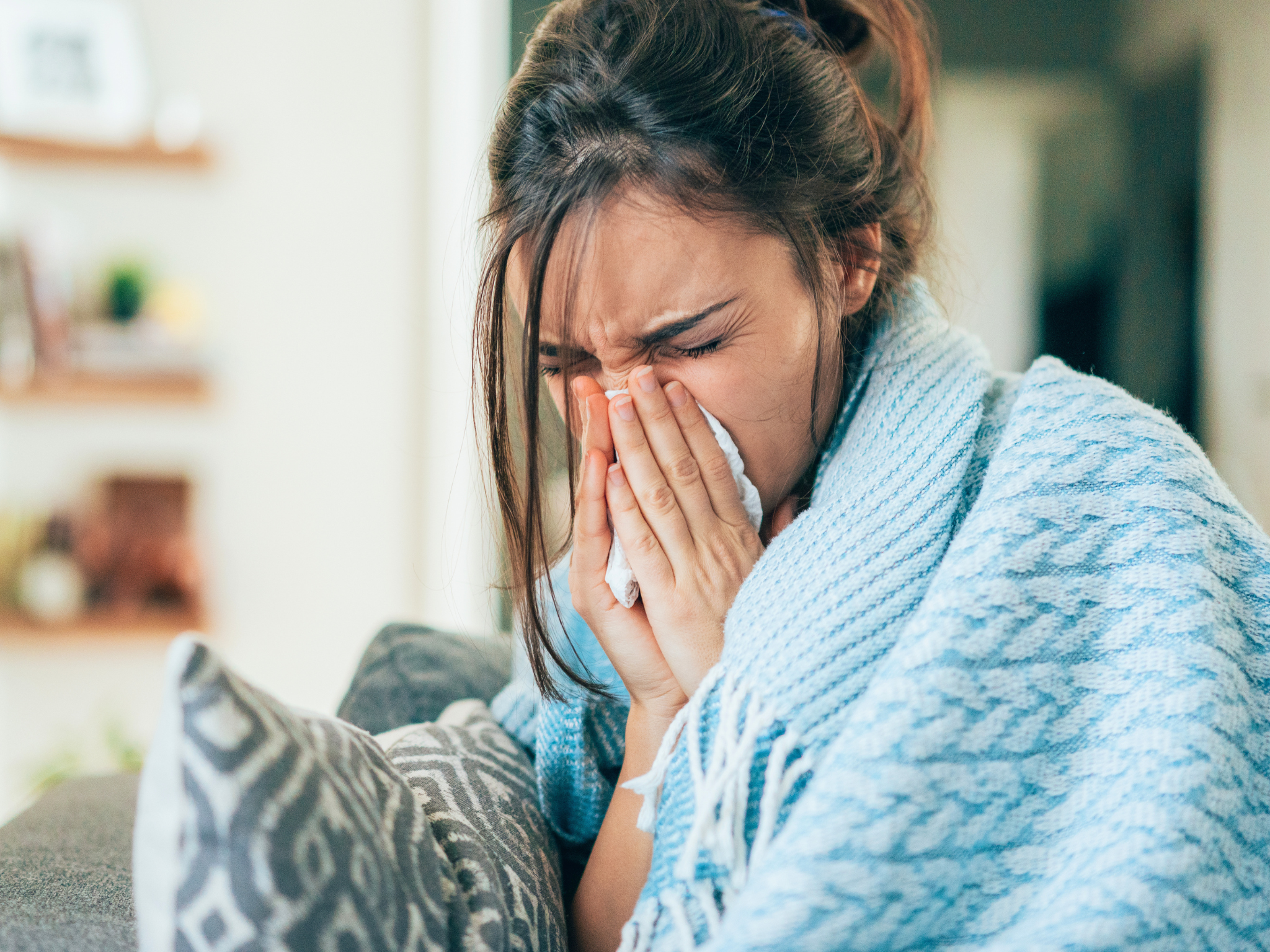 female on couch wrapped in light blue blanket blowing nose during virus season